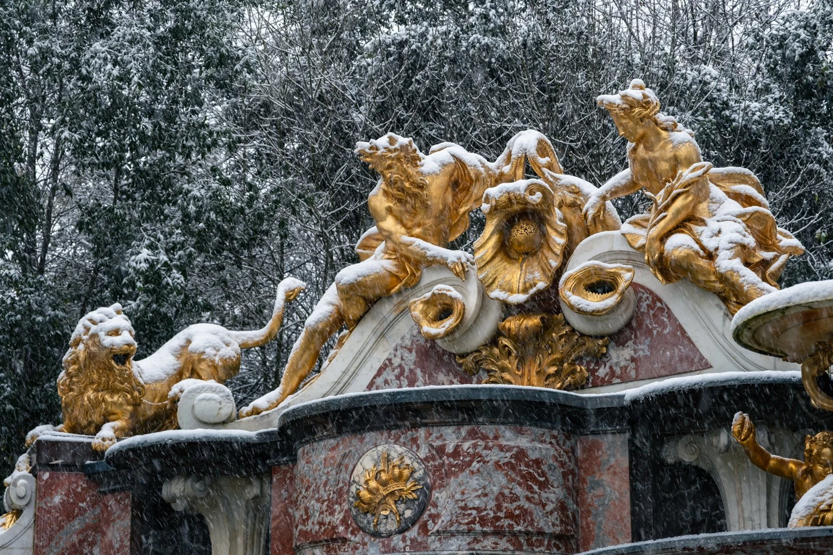 Snow-covered ornate gold and white sculpture featuring mythological animals and figures, set against a background of snow-covered trees.