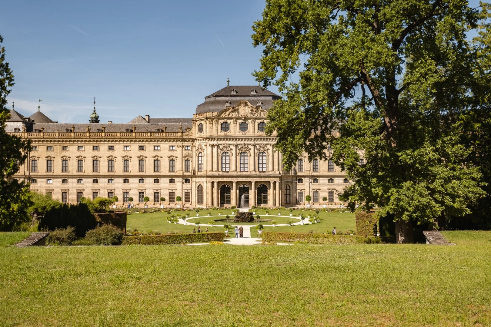 A grand historic building with ornate architecture, large windows, and a central entrance, set behind a well-maintained park with green lawn, walking paths, trimmed bushes, and tall trees under a clear blue sky.