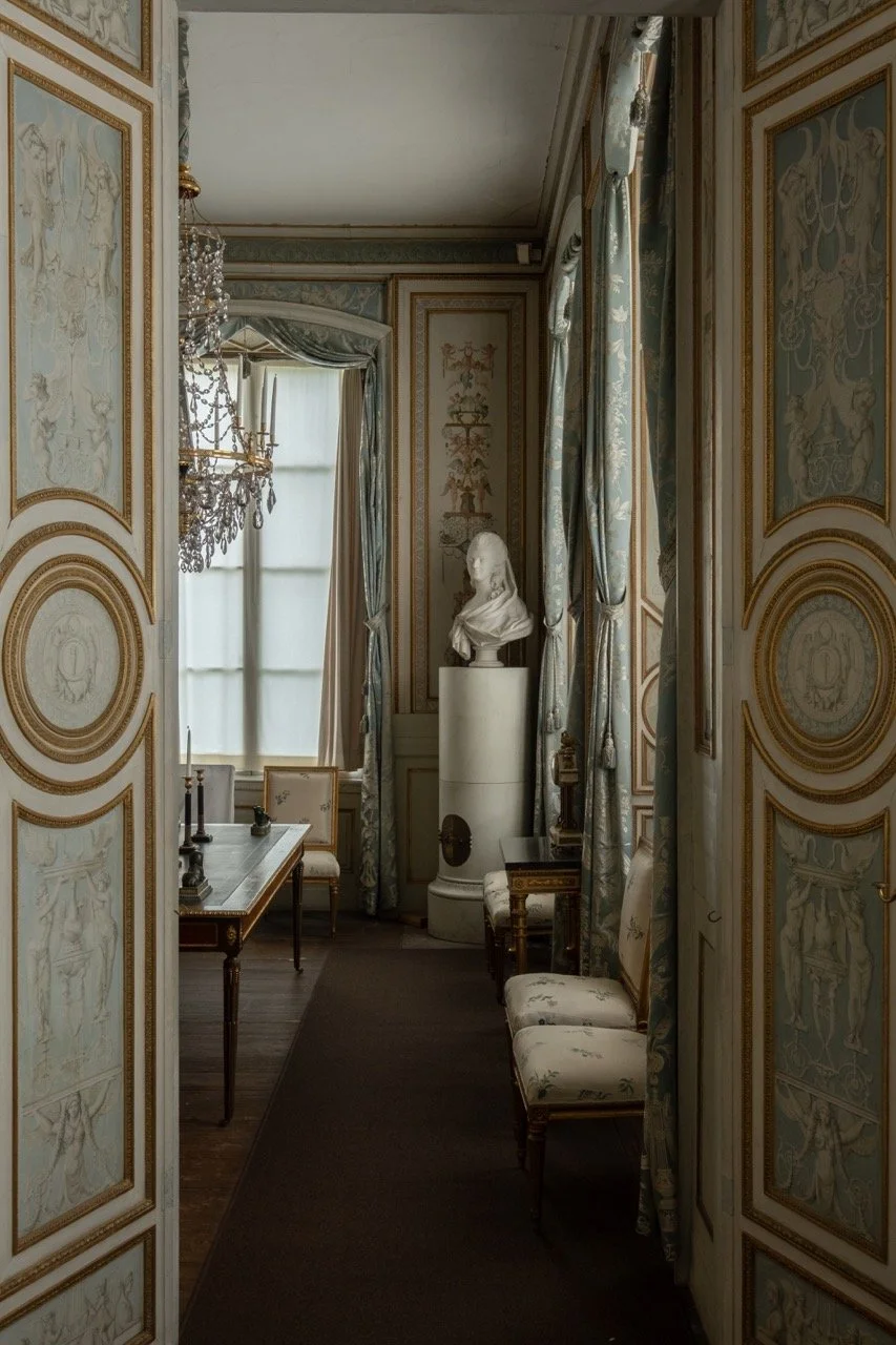 An elegant, historic room with ornate painted walls, tall windows with drapes, a chandelier, a marble bust on a pedestal, and antique furniture.