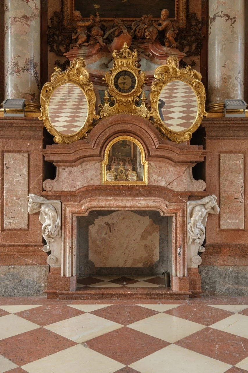 An ornate fireplace with marble columns, a gold clock, and decorative mirrors, set in a richly decorated room with marble and wooden elements.