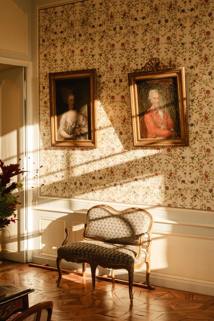 A vintage-style decorated room with floral wallpaper, two framed classic paintings hanging on the wall, a wooden chair with patterned upholstery, and sunlight streaming through a window casting shadows on the wall and furniture.