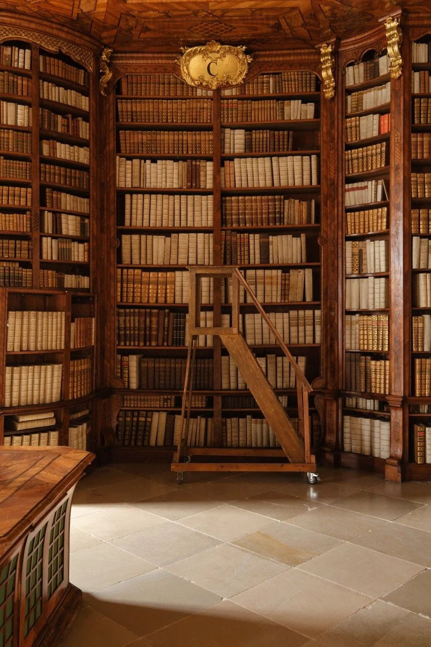 A grand wooden library with tall bookshelves filled with numerous old, leather-bound books. The shelves are ornately decorated with gold accents and carved details. There is a small, mobile wooden ladder in front of the shelves for reaching high book
