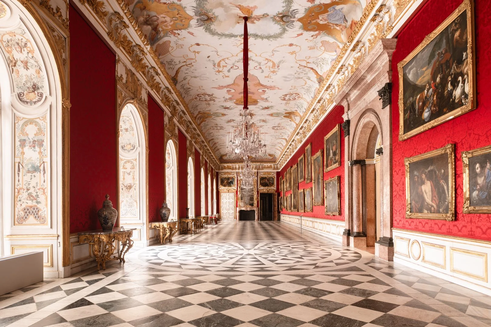Opulent museum hall with red walls adorned with paintings in gilded frames, a black-and-white checkered marble floor, ornate ceiling frescoes, and crystal chandeliers hanging from the ceiling.