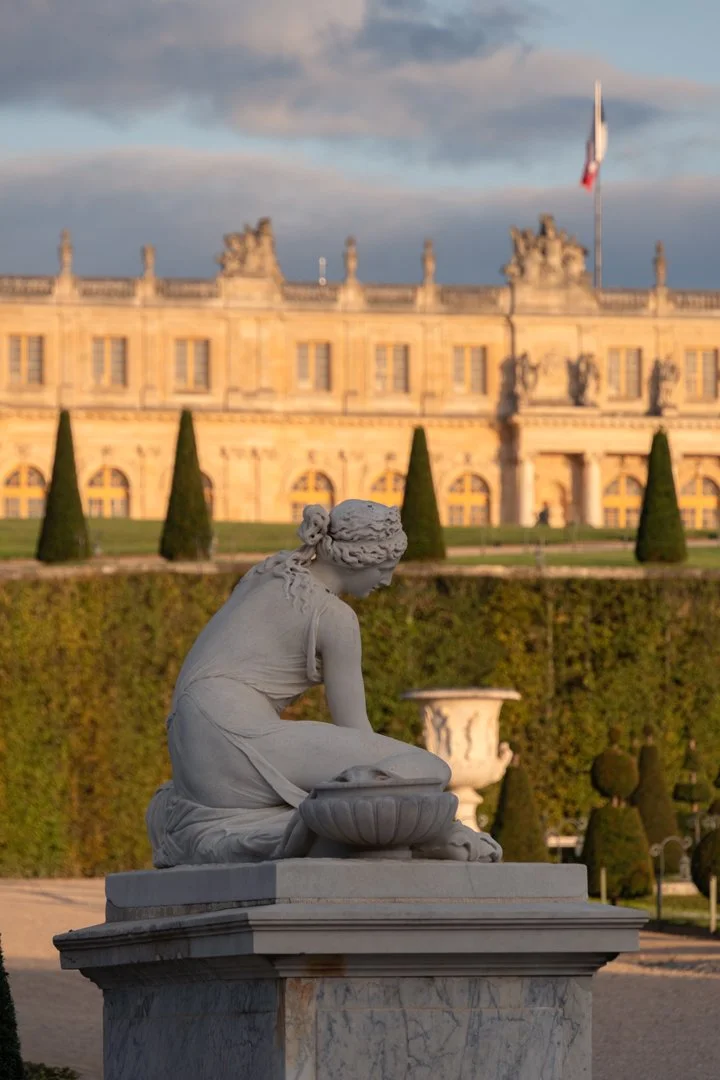 A marble statue of a seated woman in a garden with manicured bushes and trees, and a historic building with a flag on top in the background.