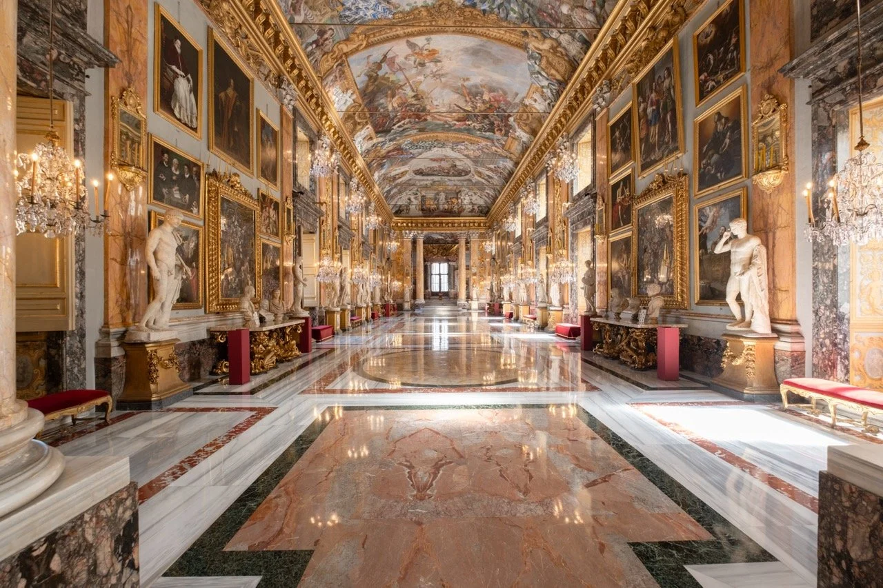 A grand, ornate hallway in a palace or museum with marble floors, gilded walls, and numerous classical statues and paintings. The ceiling is decorated with elaborate frescoes and chandeliers hang on the walls.