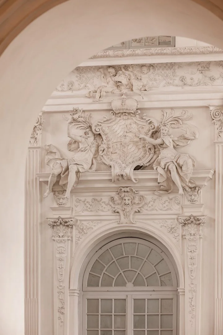 An ornate white baroque-style wall with intricate carvings and sculptures, including two seated angelic figures flanking a detailed crest or coat of arms above a window or doorway.