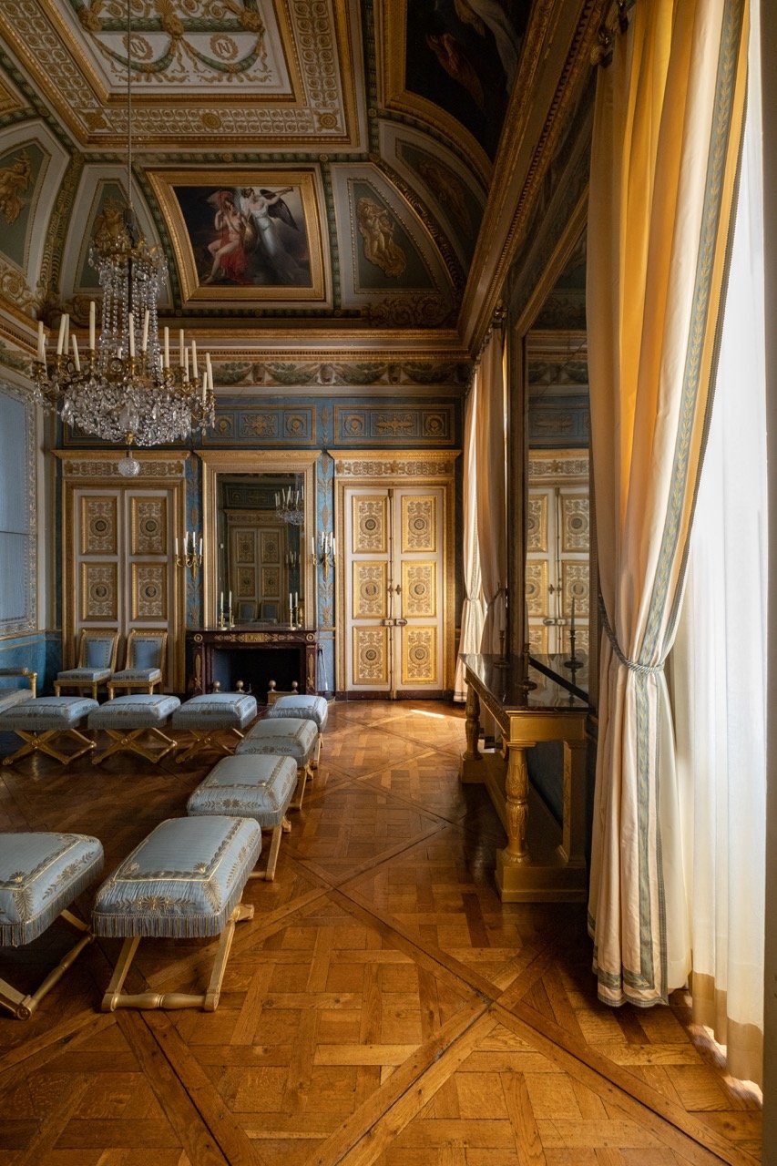 An ornate, historical ballroom with blue and gold decorative paneling, a large crystal chandelier, portraits on the ceiling, and yellow curtains on tall windows.