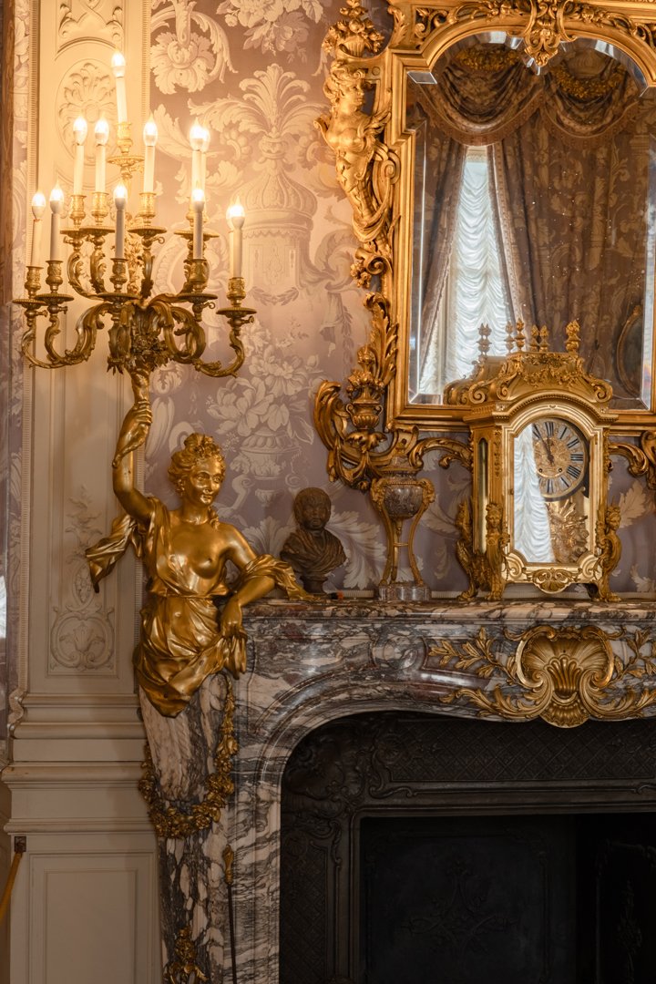 Elegant room with ornate gold-framed mirror, marble fireplace, and gold sculptures including a woman holding a candelabrum and a small bust, with luxurious curtains reflected in the mirror.