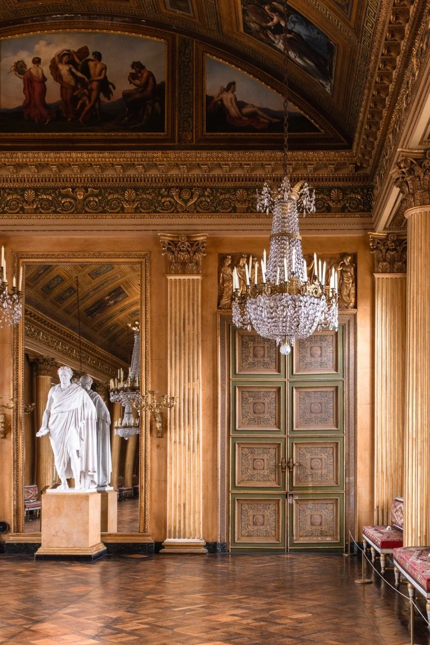 Elegant room with ornate gold and green double doors, grand crystal chandelier, marble statue, large mirror, painted ceiling, and classical decor.