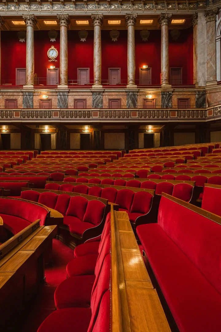 Interior of an elegant theater or opera house with red velvet seats, marble columns, and ornate architectural details.