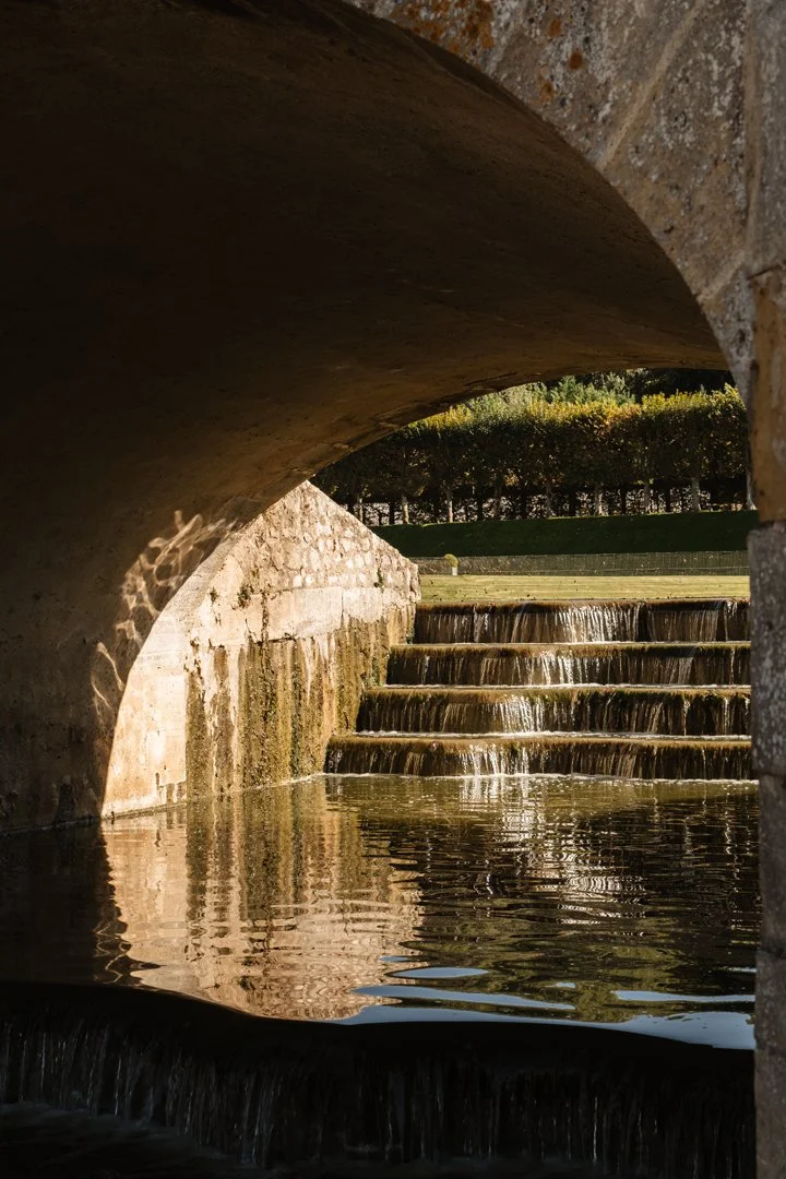 Water flowing down tiered steps under a bridge, with trees in the background.