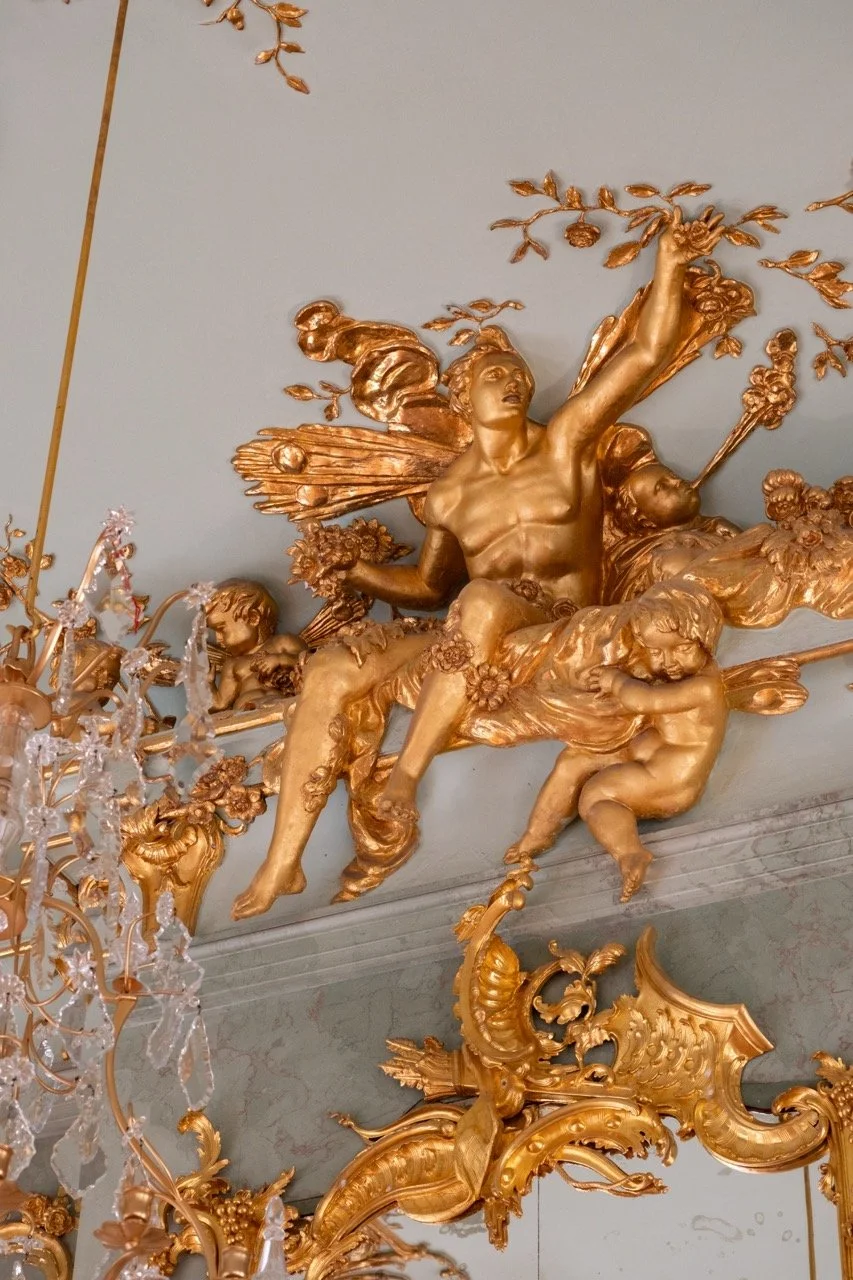 Gold ornate sculpture of mythological figures with winged women and cherubs on a ceiling, with a crystal chandelier partially visible on the left.