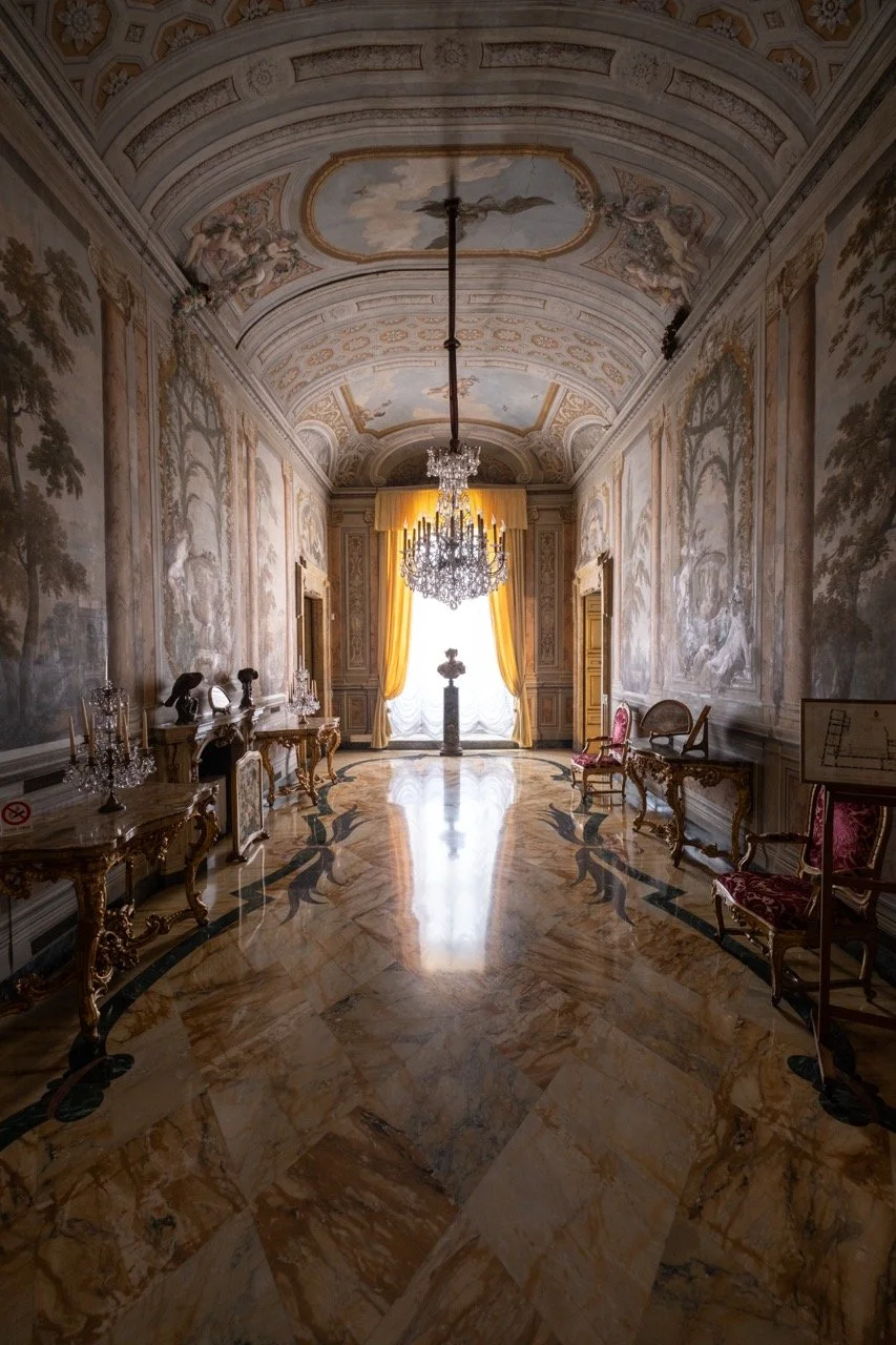 Elegant, ornate hallway with a marble floor, gold-accented furniture, large chandelier, and decorative painted walls with drapes at the end.