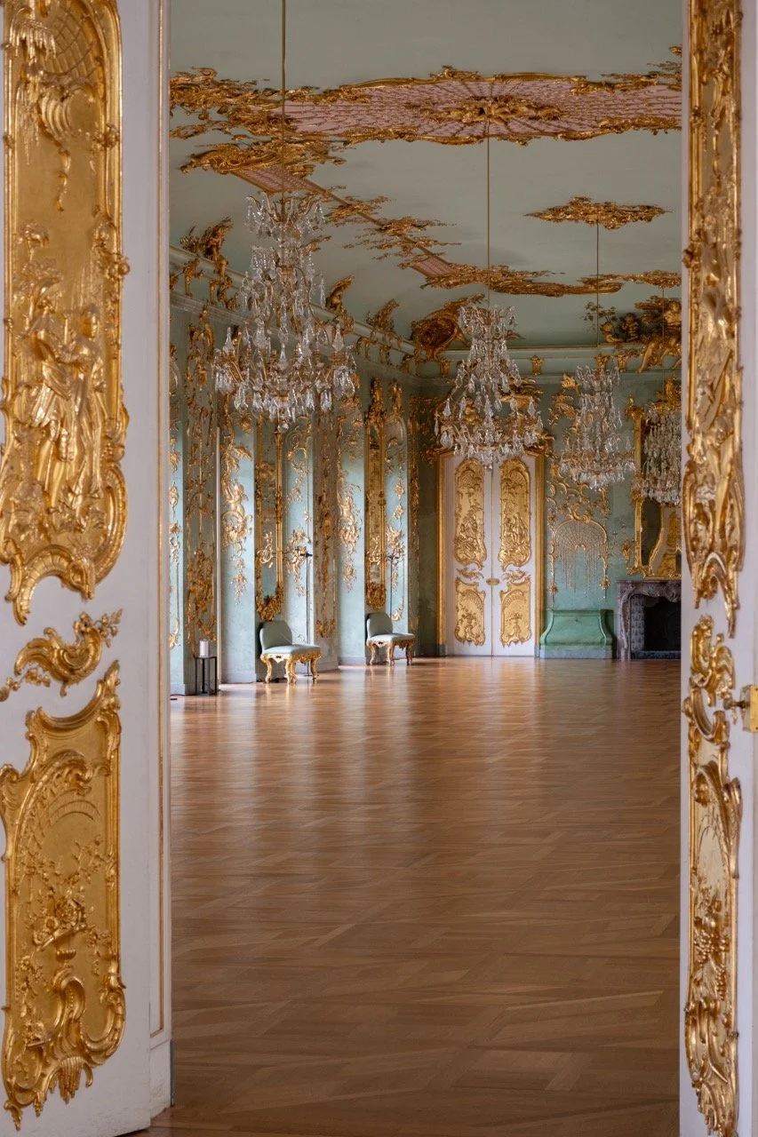 View of an elegant, ornate room with gold trim and moldings, large crystal chandeliers, tall windows, and antique furniture.