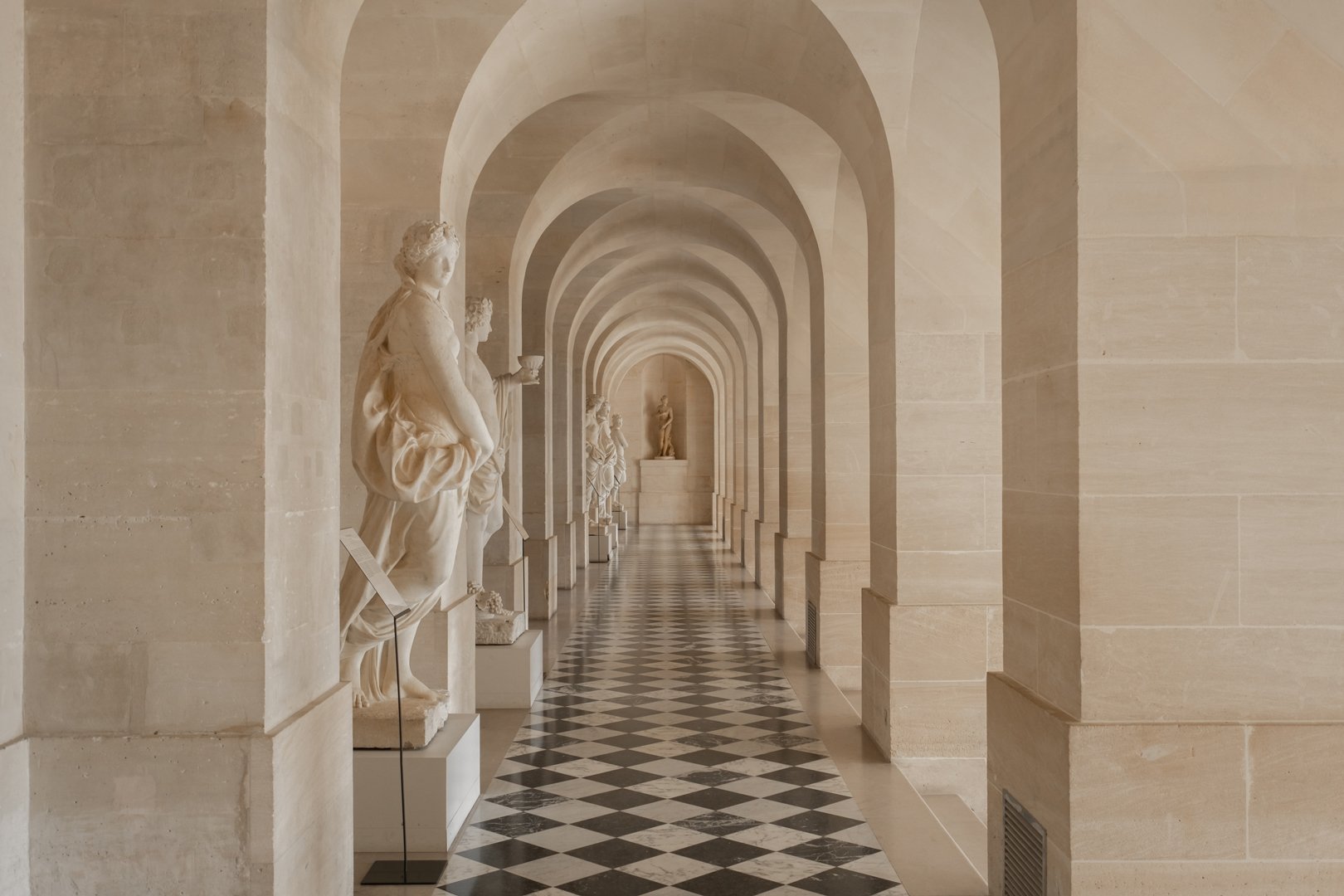 Museum corridor with beige stone walls, black and white checkered floor, and classical marble statues on white pedestals along the side.