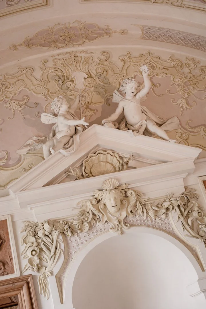 Ornate ceiling with decorative stucco sculptures, including cherubs and a female face surrounded by floral and shell motifs.