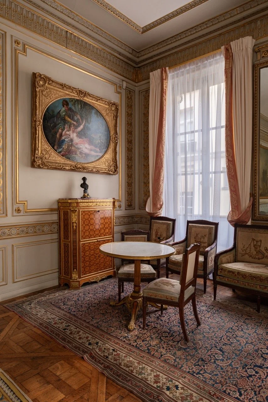 Elegant vintage room with ornate gold-trimmed walls, a large window with curtains, a round marble-topped table, antique chairs with floral upholstery, a decorative wooden cabinet, a framed painting, a Persian rug, and a small sculpture on the cabinet