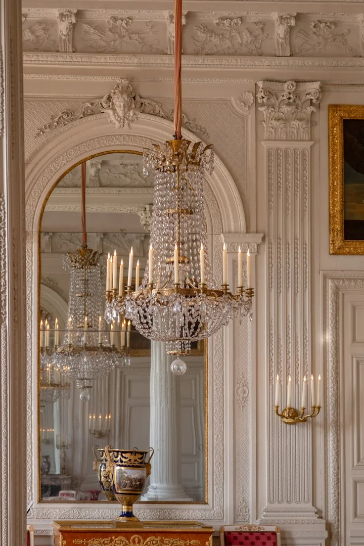 A luxurious European-style room with ornate white molding, a large mirror, and elegant gold and crystal chandeliers with lit candles. There's also a decorative urn on a table and a red upholstered chair.