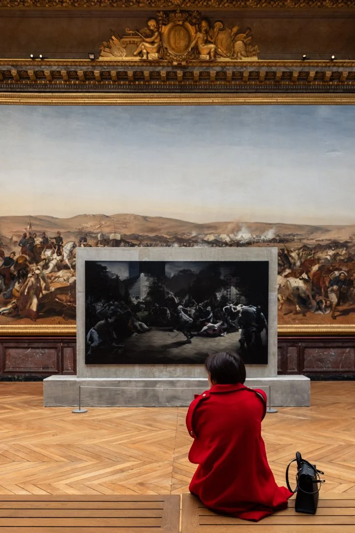 A woman in a red coat sitting on the floor in an art museum, facing a framed painting depicting a historical battle scene with soldiers and horses, with a large landscape mural behind it.