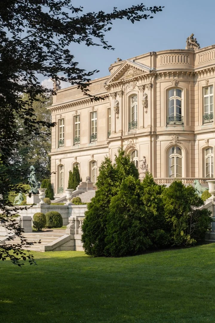 A grand, ornate mansion with classical architecture, balcony railings, and intricate decorative details. Surrounded by well-manicured lawns, shrubs, and trees.