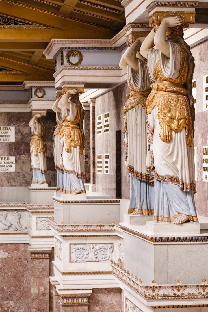 Series of ancient Greek-style statues with lifted arms, draped clothing, and lion-head belts, displayed on ornate pedestals inside a decorated building.
