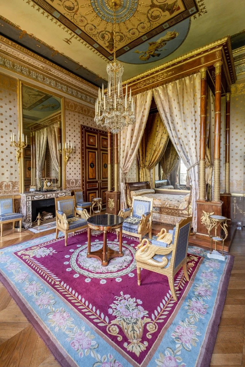 An ornate, luxurious room with a chandelier hanging from a decorated ceiling, gold accents, a mirror above a marble fireplace, and a four-poster bed with drapes. There is a red floral area rug, with a small round black table and several blue and gold