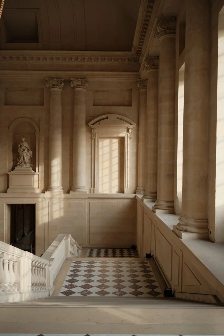 Interior of a grand, classical building with tall columns, statues, and checkered marble flooring, illuminated by sunlight through large windows.