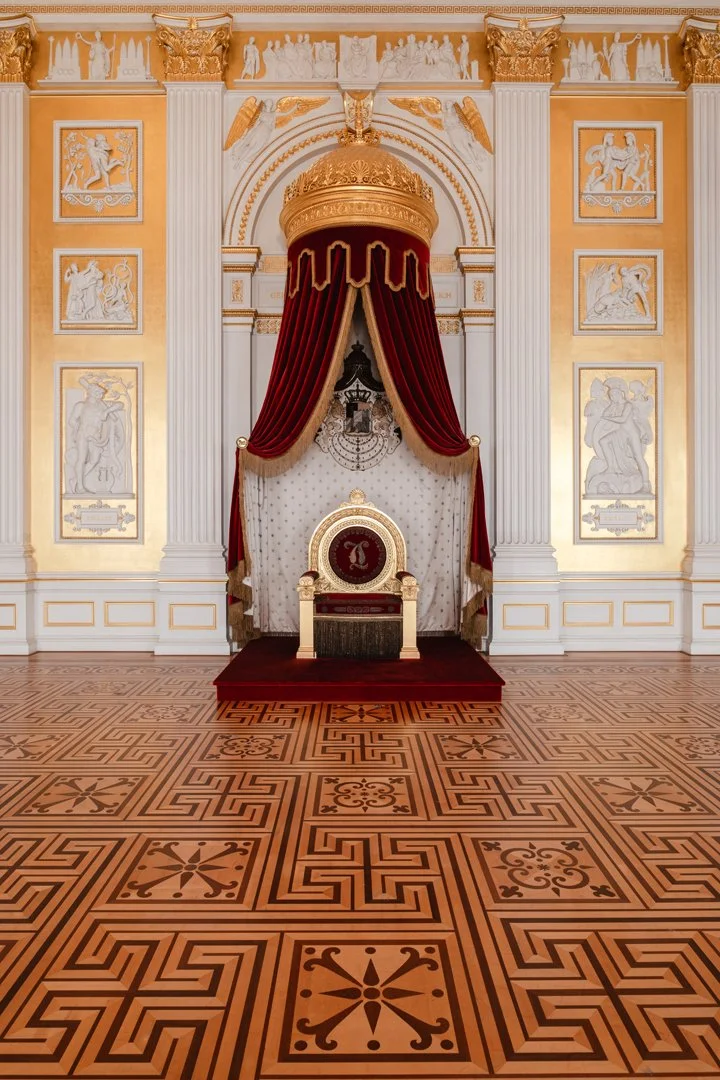 Royal throne room with a golden throne on a red carpeted platform, ornate drapery, and classical wall decorations in gold and white.