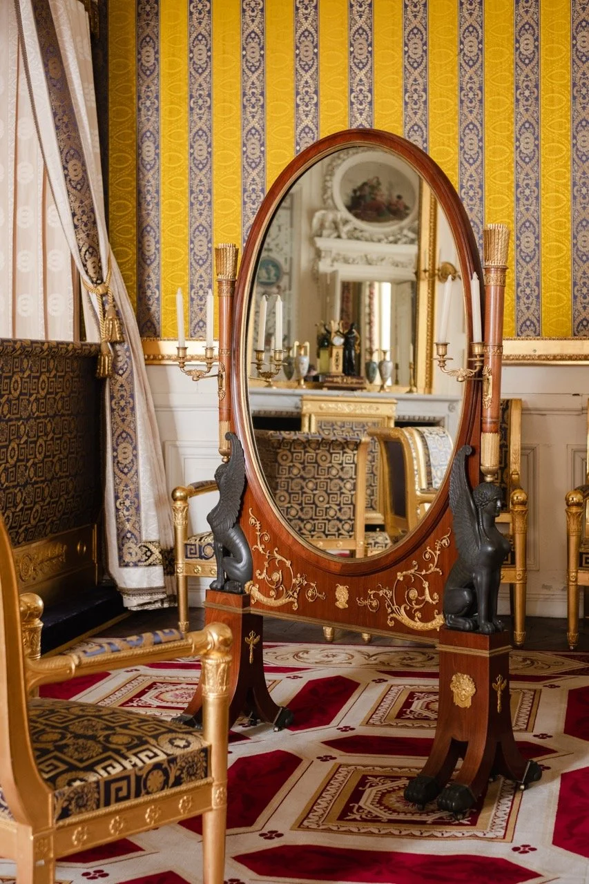 An ornate, vintage room with gold accents, patterned curtains, and a large wooden oval mirror on a stand with decorative black griffins. The room has patterned wallpaper, matching chairs, and a red, gold, and cream carpet.