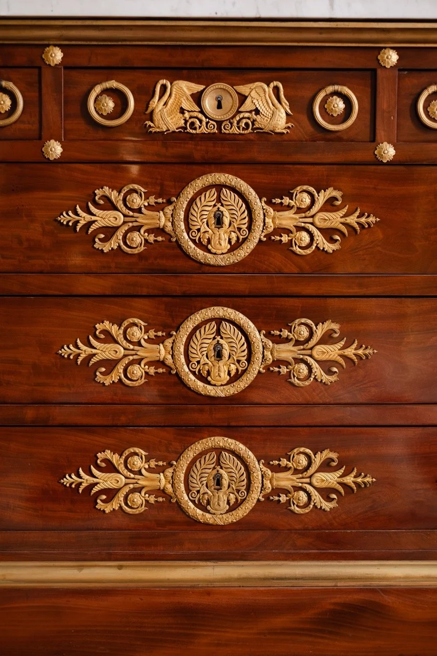 Three ornate wooden drawer handles with gold-colored floral and scroll designs on a polished wooden surface, with a decorative crest above each handle.