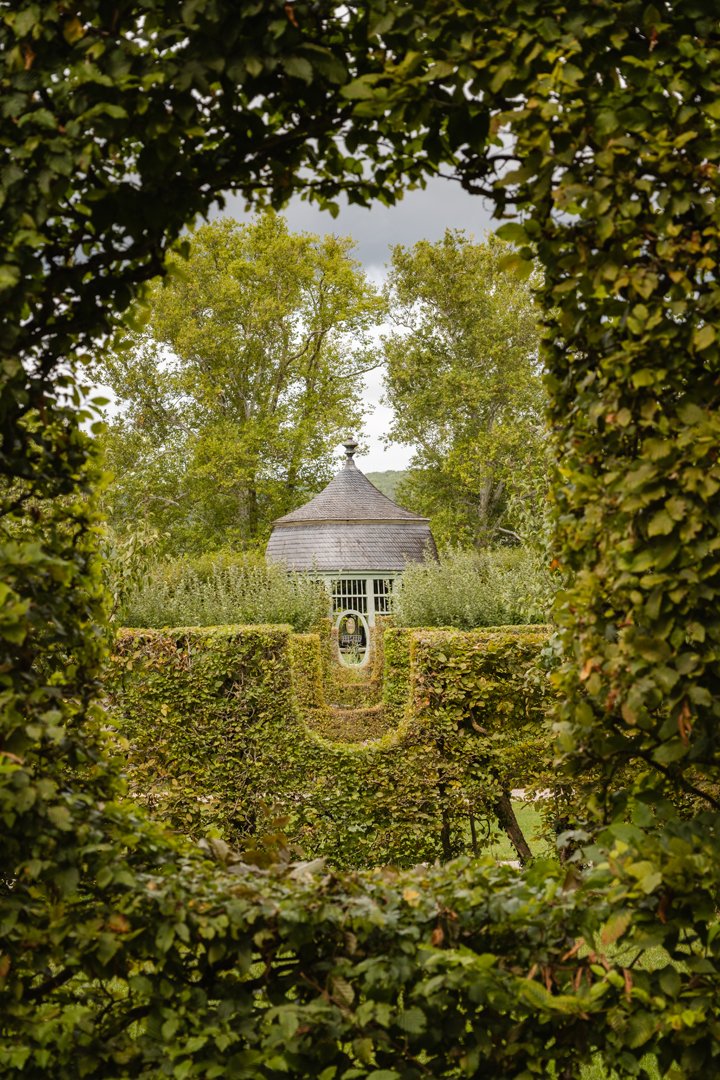 A view through a hedge framing a garden with a gazebo in the distance, surrounded by trees.