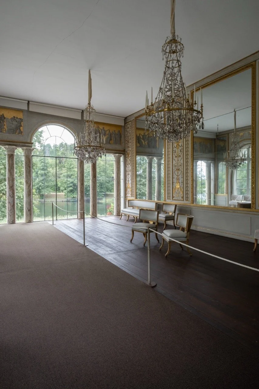 Elegant room with large glass windows, ornate gold and white decor, chandeliers, and vintage seating.