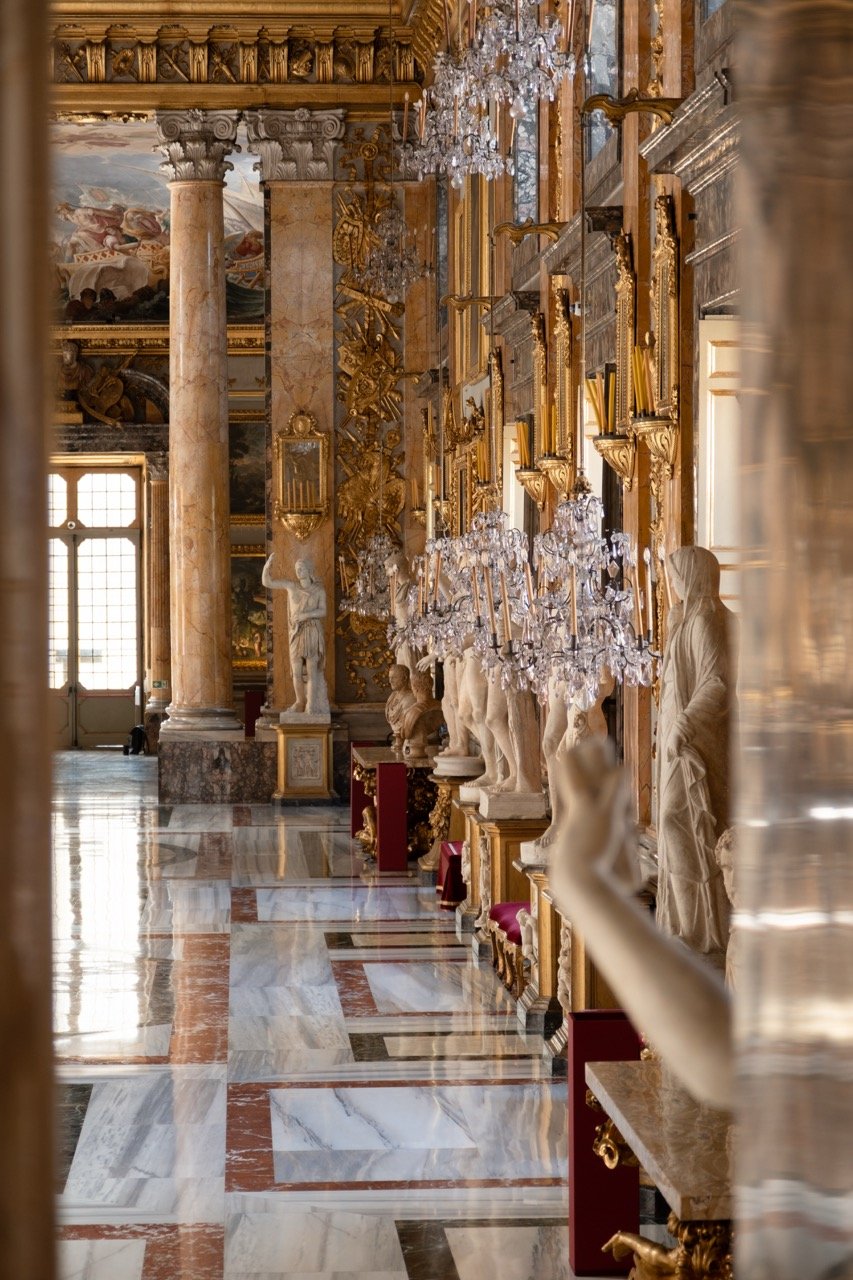 An ornate, gilded hall with marble floors, classical statues, and decorative chandeliers. Large windows allow natural light to illuminate the space.