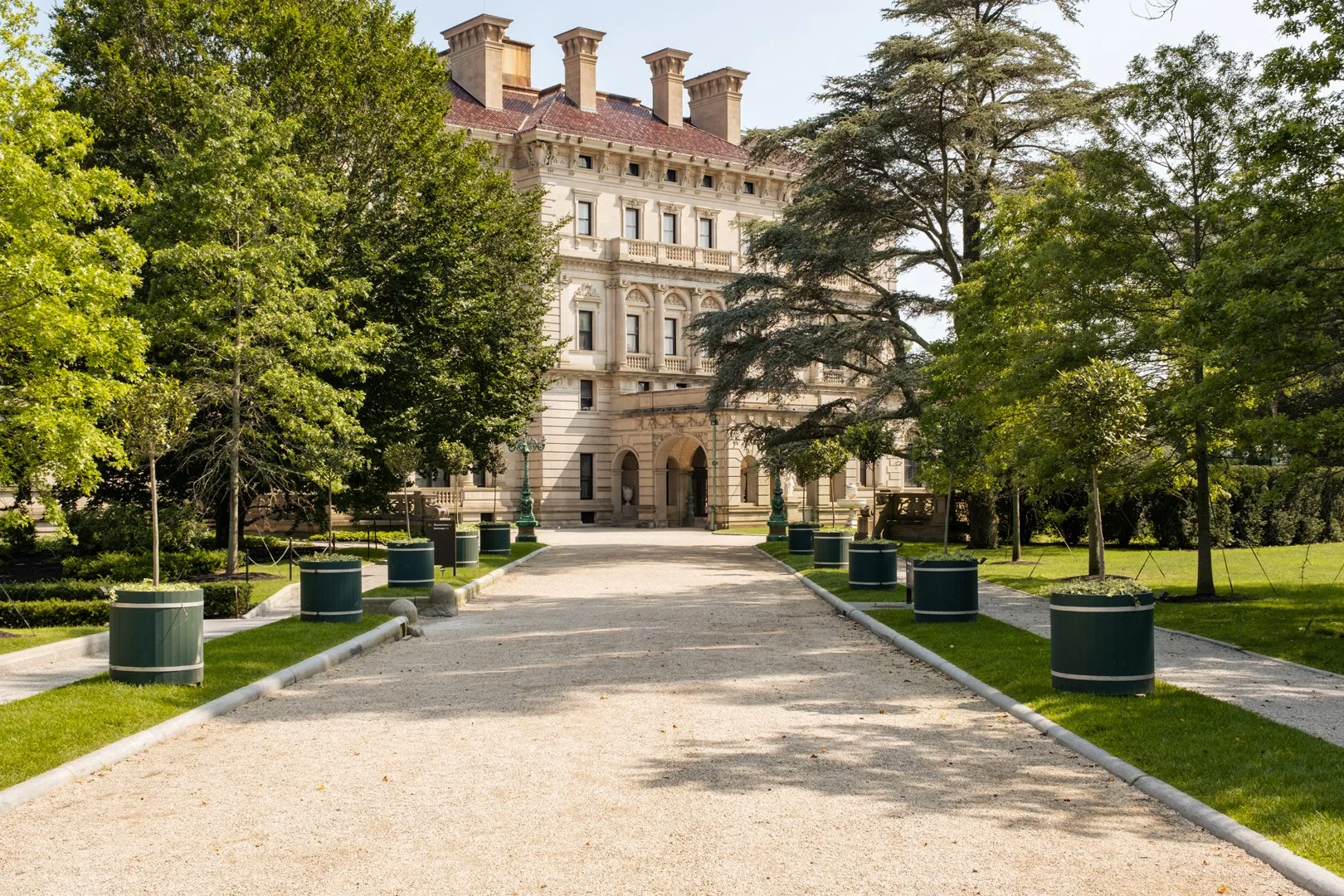 A grand, historic building with a red tile roof and multiple chimneys, framed by lush green trees, with a gravel pathway leading up to the entrance.