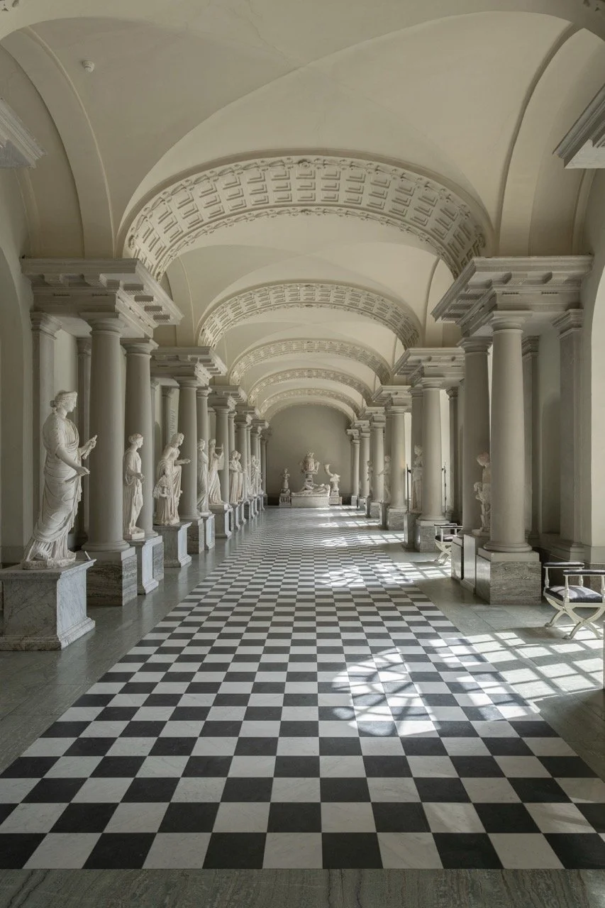 A grand hall featuring marble statues on pedestals, tall columns, vaulted ceilings, and a inlaid black-and-white checkered floor.