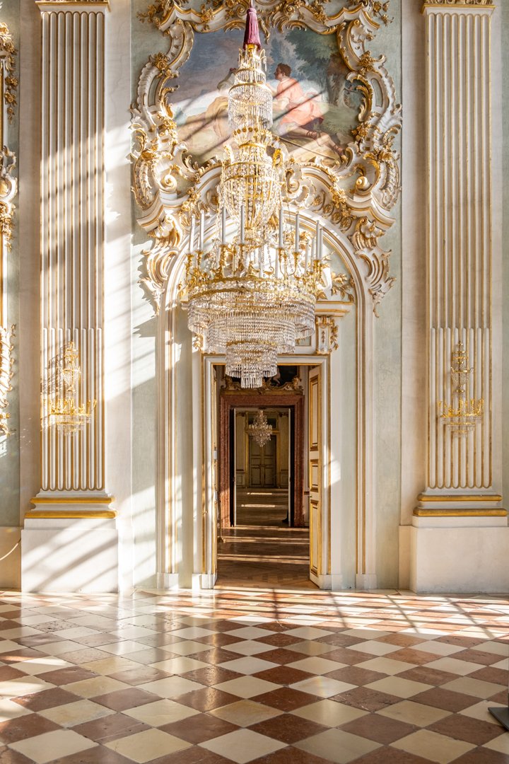 An ornate room with gilded architectural details, large chandeliers, and a checkered marble floor. Light streams in through windows, casting shadows on the walls and floor. A doorway leads into another similarly decorated room.