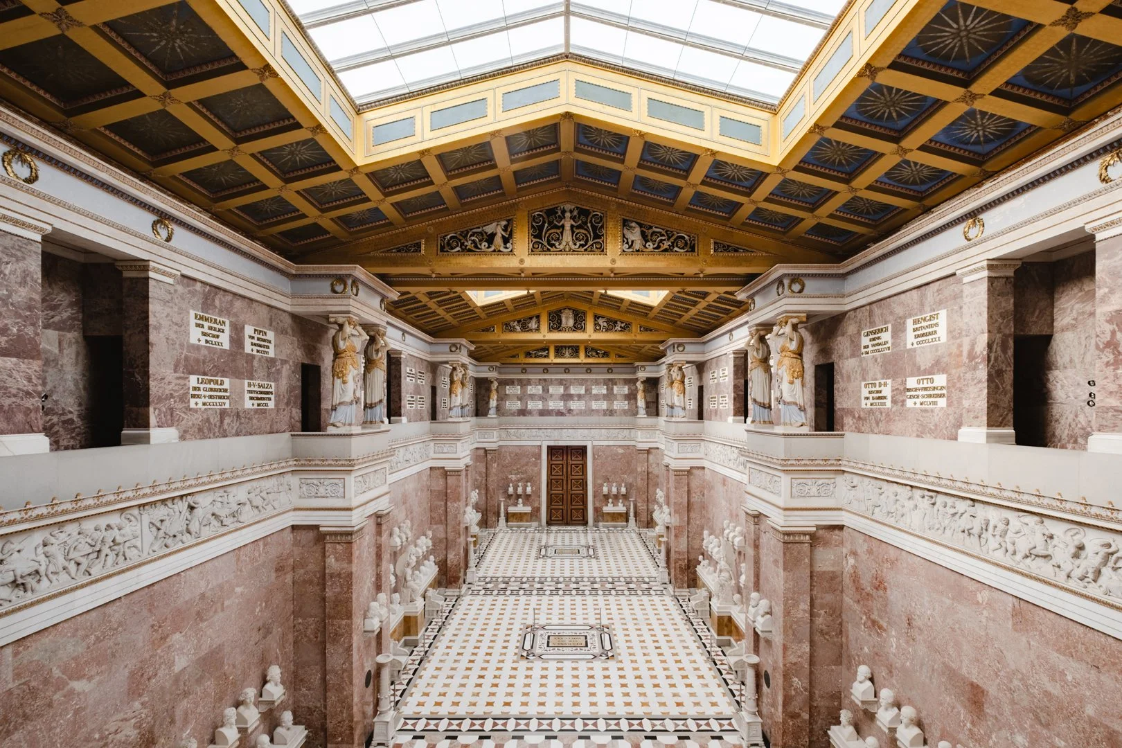 Interior of a grand museum hall with marble walls, decorative sculptures, and a glass skylight ceiling.