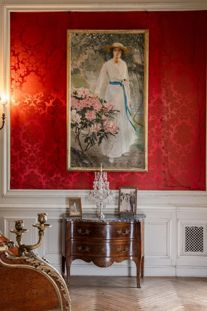Interior of an elegant room with red wallpaper, a large framed painting of a woman in a white dress and hat, an antique wooden dresser with a marble top, a crystal chandelier, and framed black-and-white photographs.