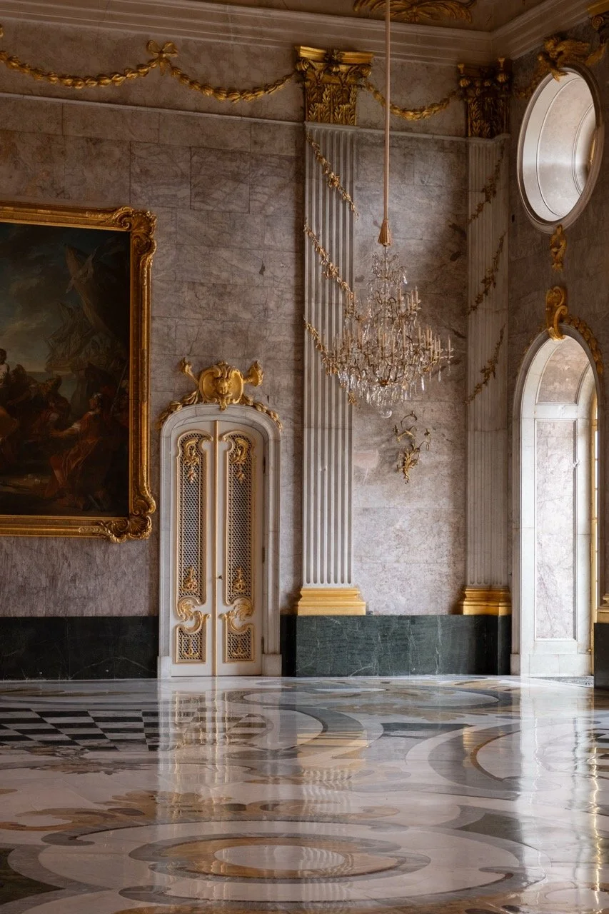 Luxurious marble hall with ornate gold details, chandelier, large window, and classical artwork.