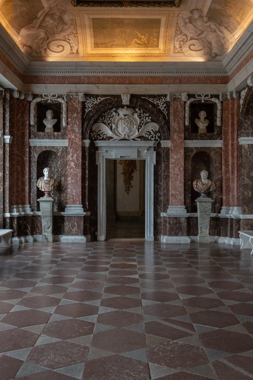 Elegant interior room with red and white marble checkered floor, ornate ceiling with fresco painting, and classical bust sculptures in niches along the walls.