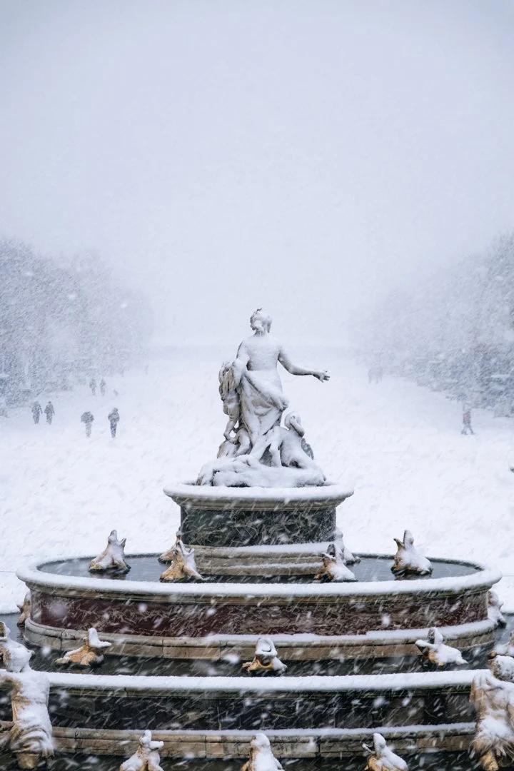 A snow-covered ornate fountain featuring a classical statue of a woman with water animals around the base, in a park during a snowstorm.