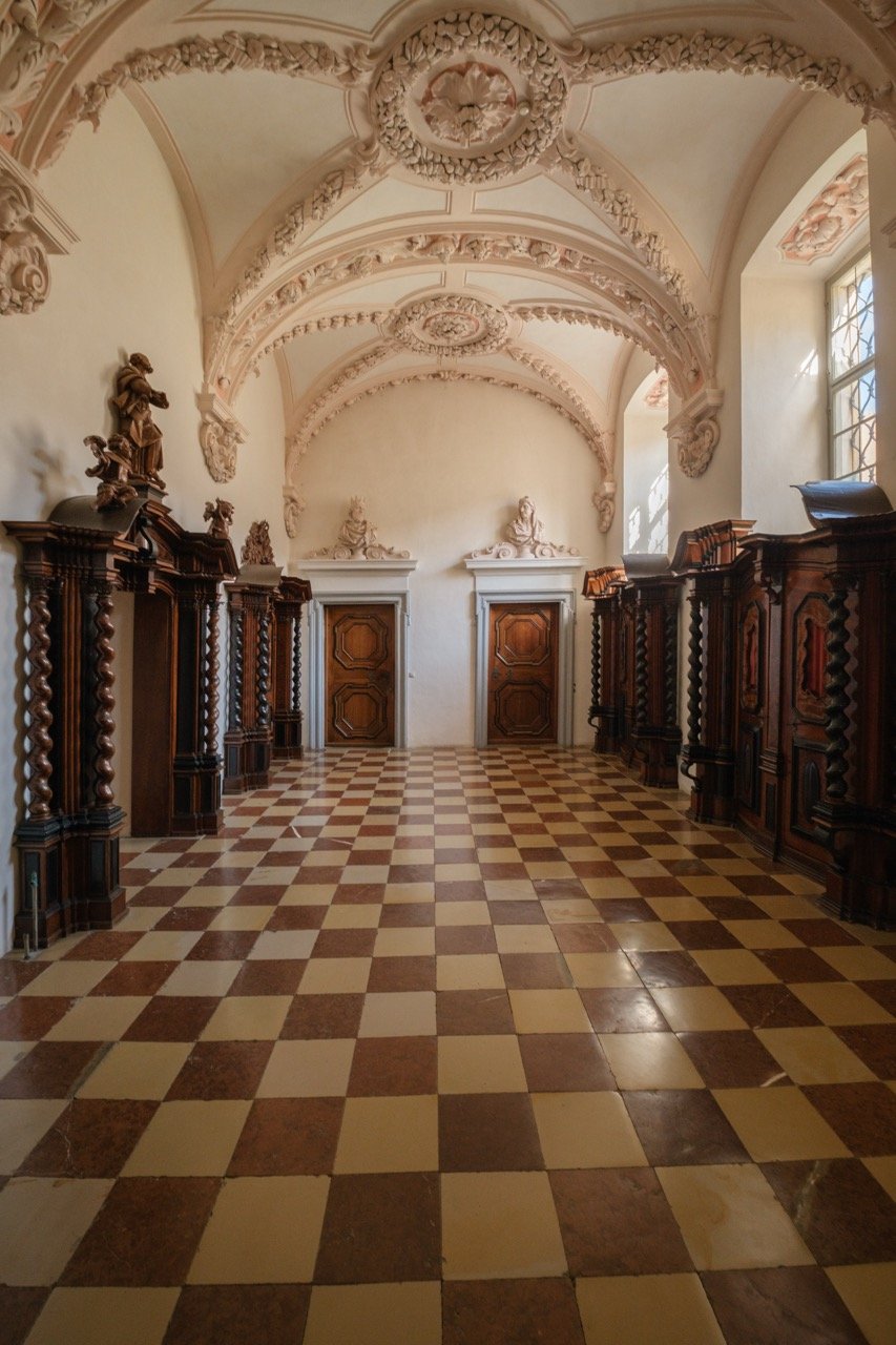 Historical room with ornate wooden furniture, carved wooden statues, decorative ceiling with intricate moldings, checkered tile floor, and large windows letting in natural light.