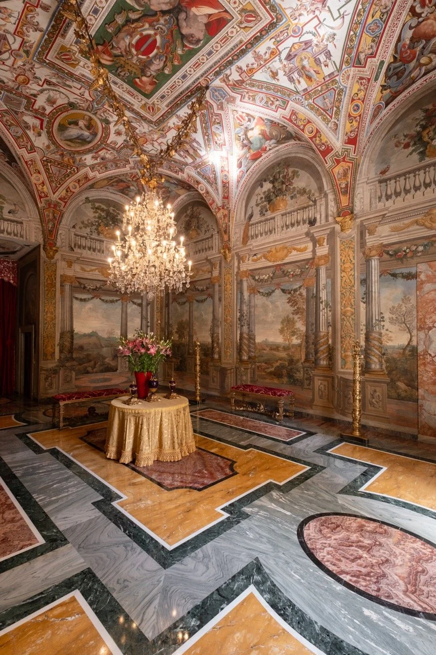 Luxurious room with elaborate painted ceilings, a chandelier, and ornate wall murals depicting landscape scenes. A round table with a gold cloth holds pink flowers in a vase, and decorative benches are along the walls.