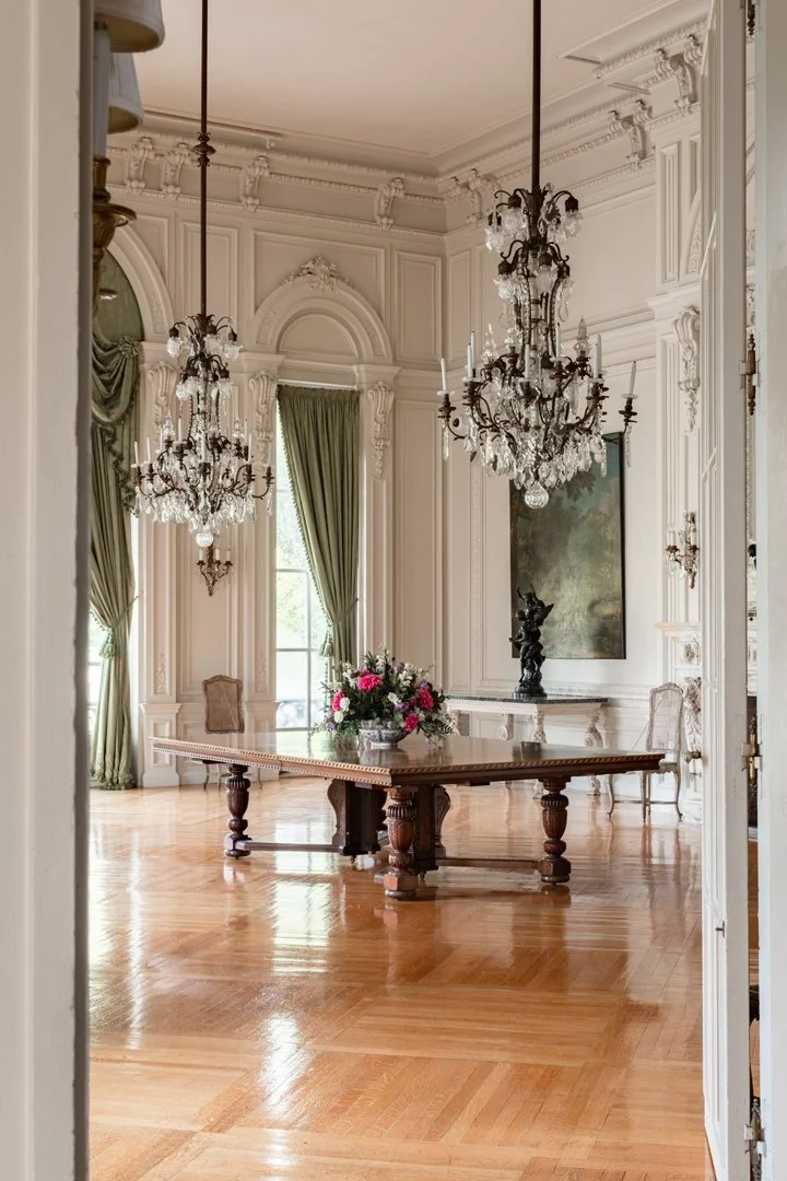 A luxurious dining room with ornate white walls, elaborate crown molding, and tall windows draped with green curtains. There are two large chandeliers hanging from the ceiling, illuminating the room, and a wooden table with a floral centerpiece. The 