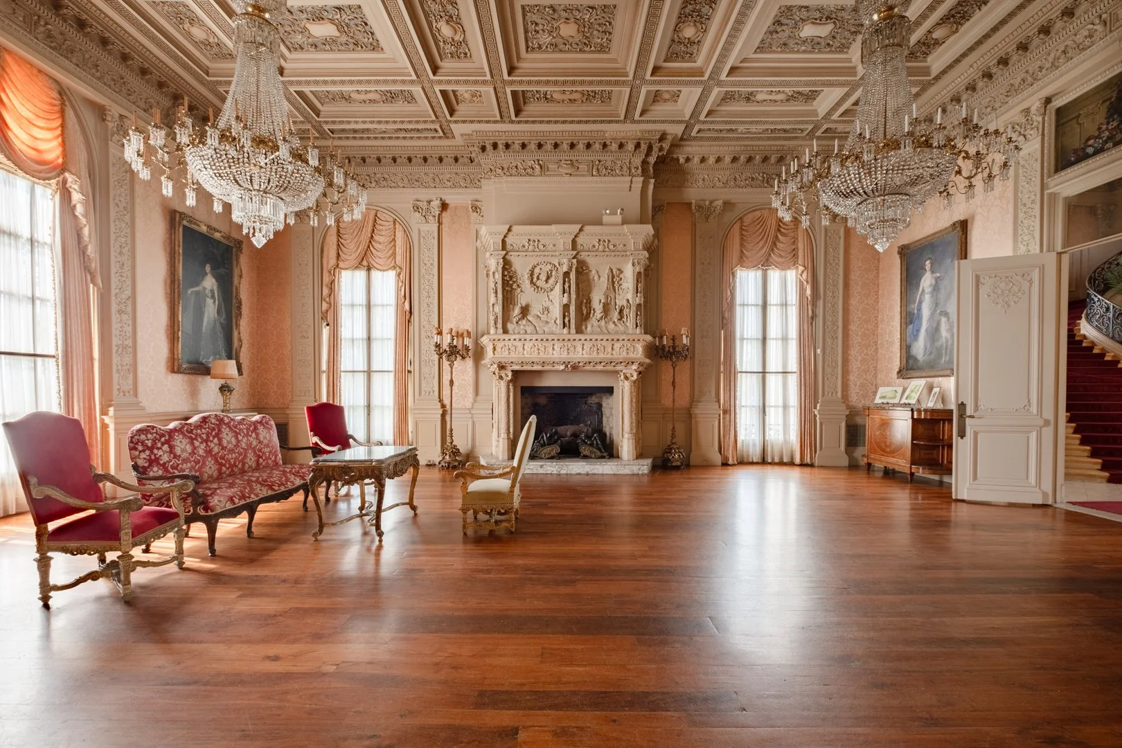 An elegant, spacious living room with ornate ceiling, crystal chandeliers, large windows with draped curtains, a marble fireplace with detailed carvings, and antique furniture including upholstered chairs and a sofa.