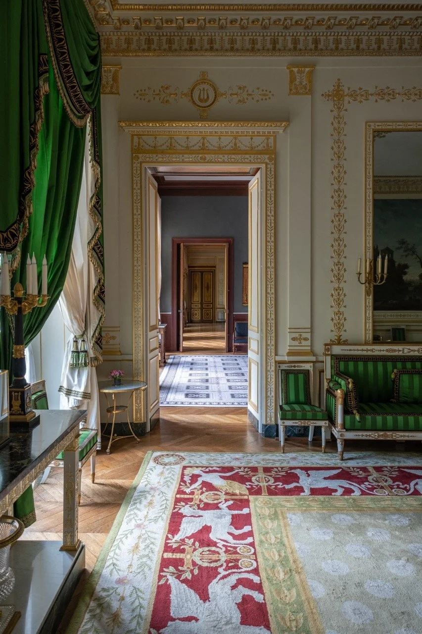 View of an ornate, luxurious room with green and gold drapes, gold-accented walls, a colorful rug with heraldic lions, and a doorway leading to more elegant, decorated rooms.