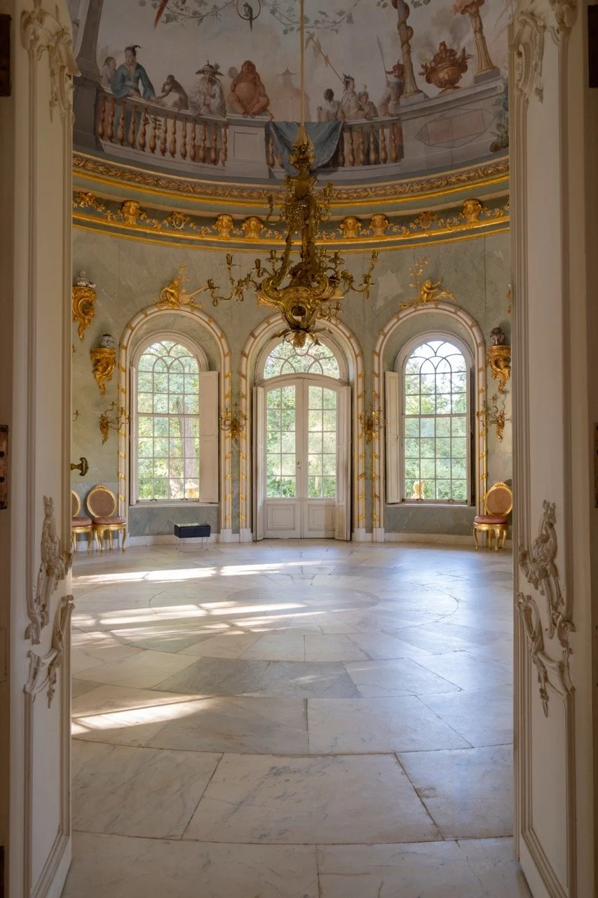 An elegant, ornate room with large arched windows, gold decorative accents, and a chandelier. The ceiling features murals, and sunlight streams onto the marble floor.