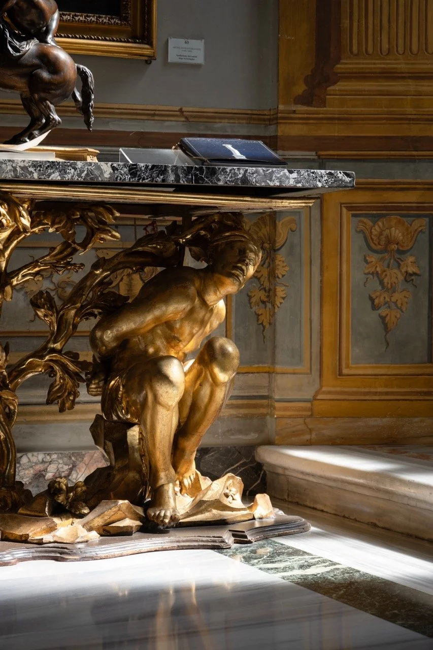 A gilded sculpture of a crouching man on a decorated table with a black marble top, located in a richly decorated room with ornate wood and gold-accented walls.