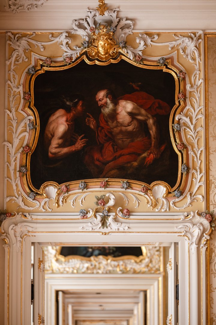 An ornate, gold-trimmed frame surrounding a dark painting of two muscular, bearded men engaged in conversation, set in a luxurious room with elaborate gold and white decor.
