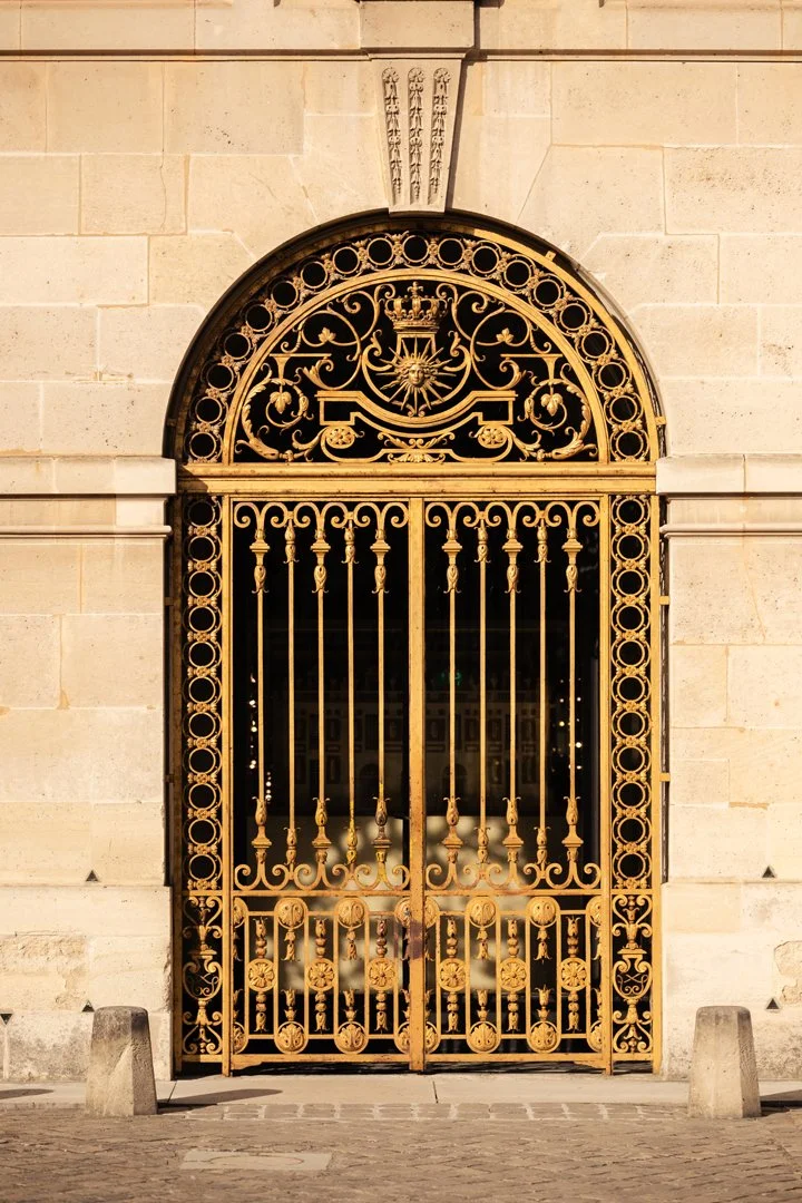 Golden decorative iron gate set in a stone wall, with ornate patterns and a crown and sun symbol at the top center.